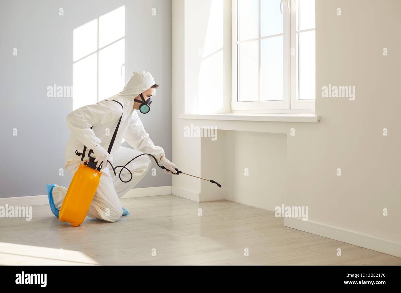 Professional exterminator in protective suit spraying insecticide in empty room for disinfection. Stock Photo