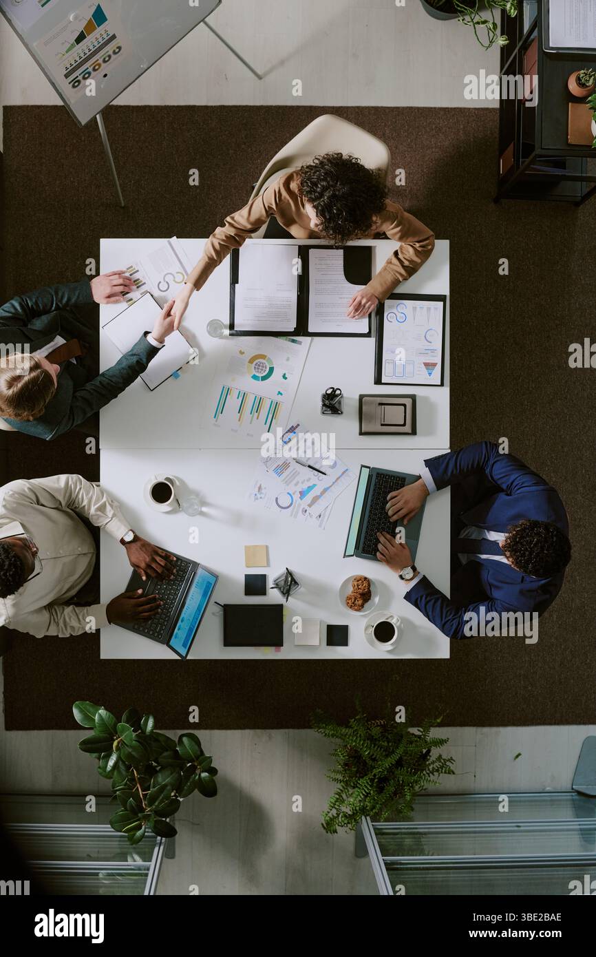 Four professionals sitting at office table, discussing work using laptops and documents. Top down view provides perspective of collaborative workspace Stock Photo