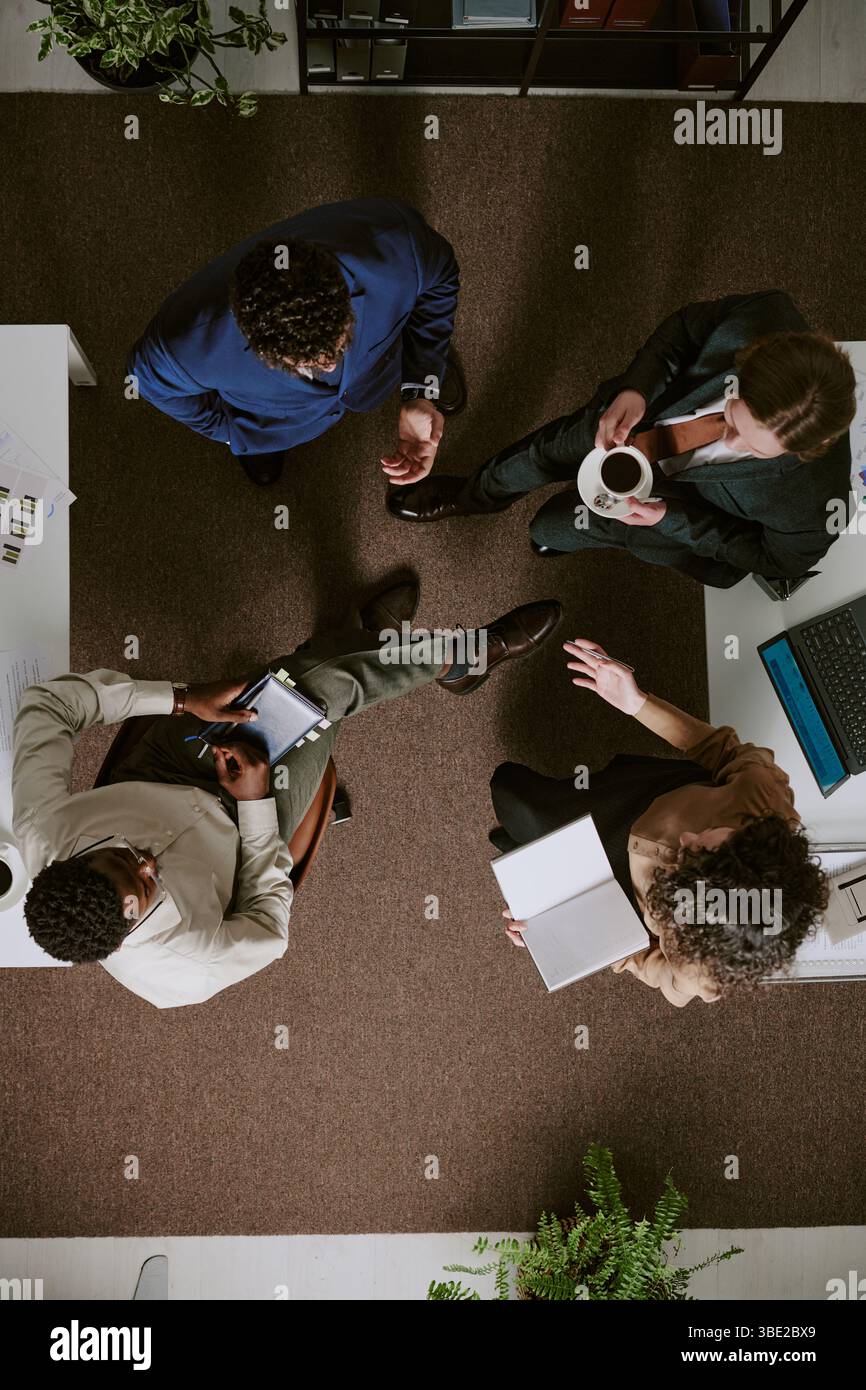 Overhead view of diverse professionals standing in office having discussion. Colleagues holding documents and coffee cups while engaging in conversati Stock Photo