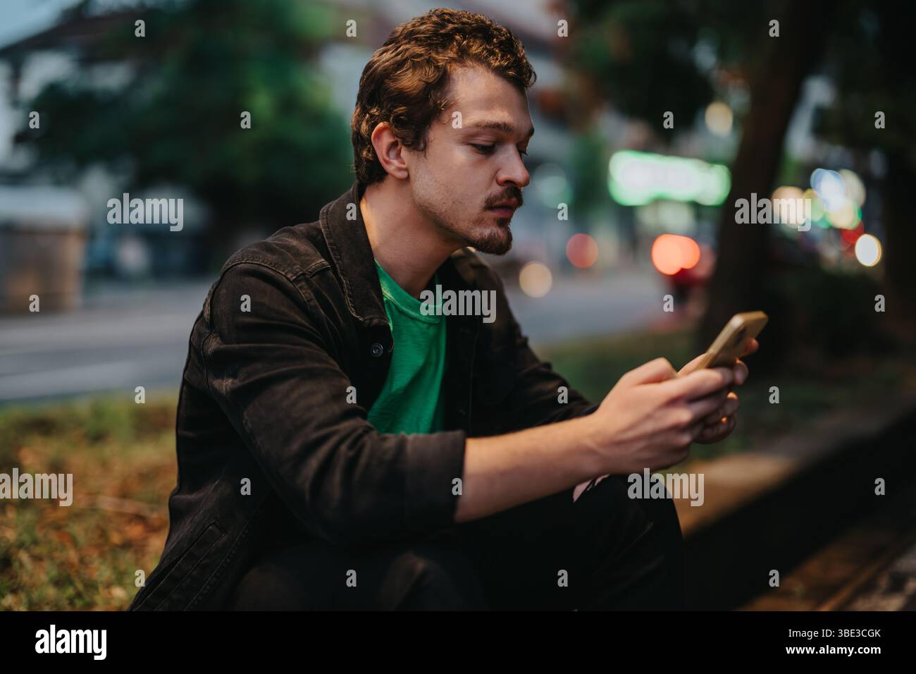 Young male sitting outdoors, looking at a smart phone during dusk Stock Photo