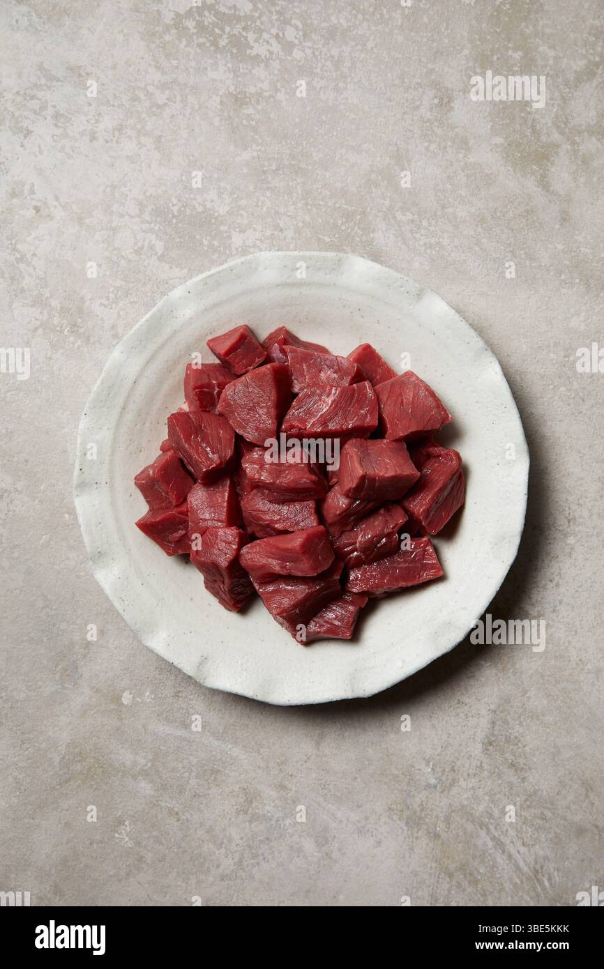 Freshly diced beef arranged on a ceramic white plate against a rustic, textured background. Perfect for depicting artisanal butchery or gourmet home c Stock Photo