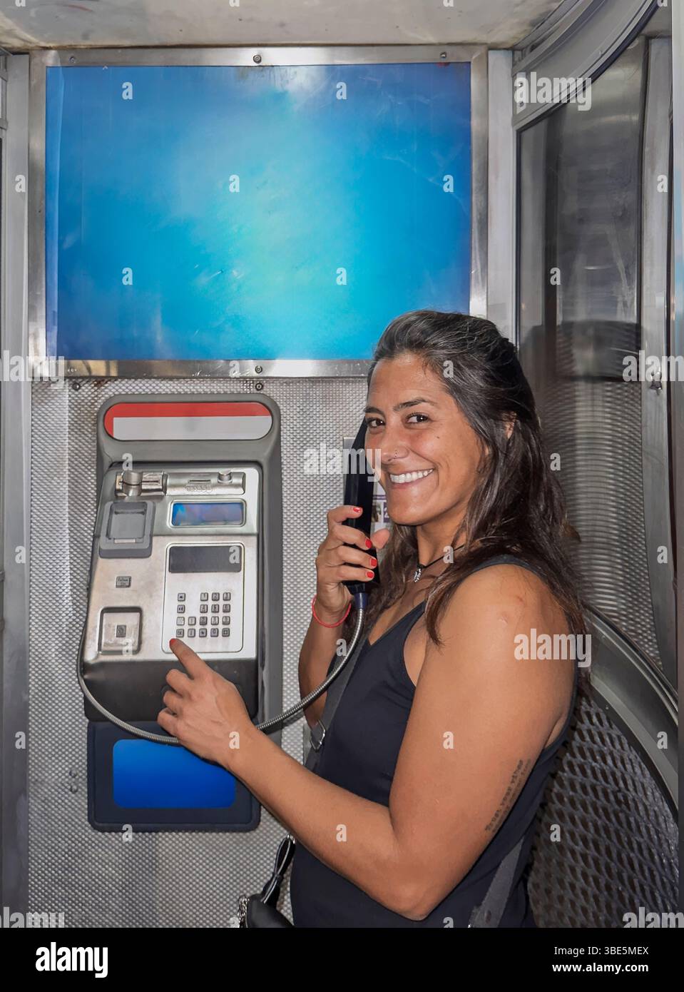A smiling woman is using a public payphone inside a metallic booth ...