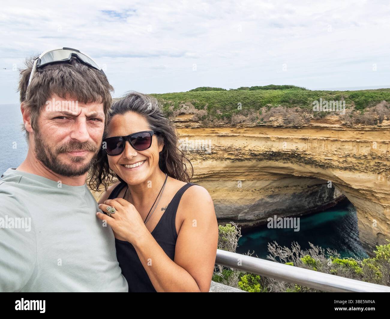 Two people smiles at the camera, enjoying a bright day by the ocean in the Twelve Apostles, Australia. They stand in front of a rugged cliff formation Stock Photo