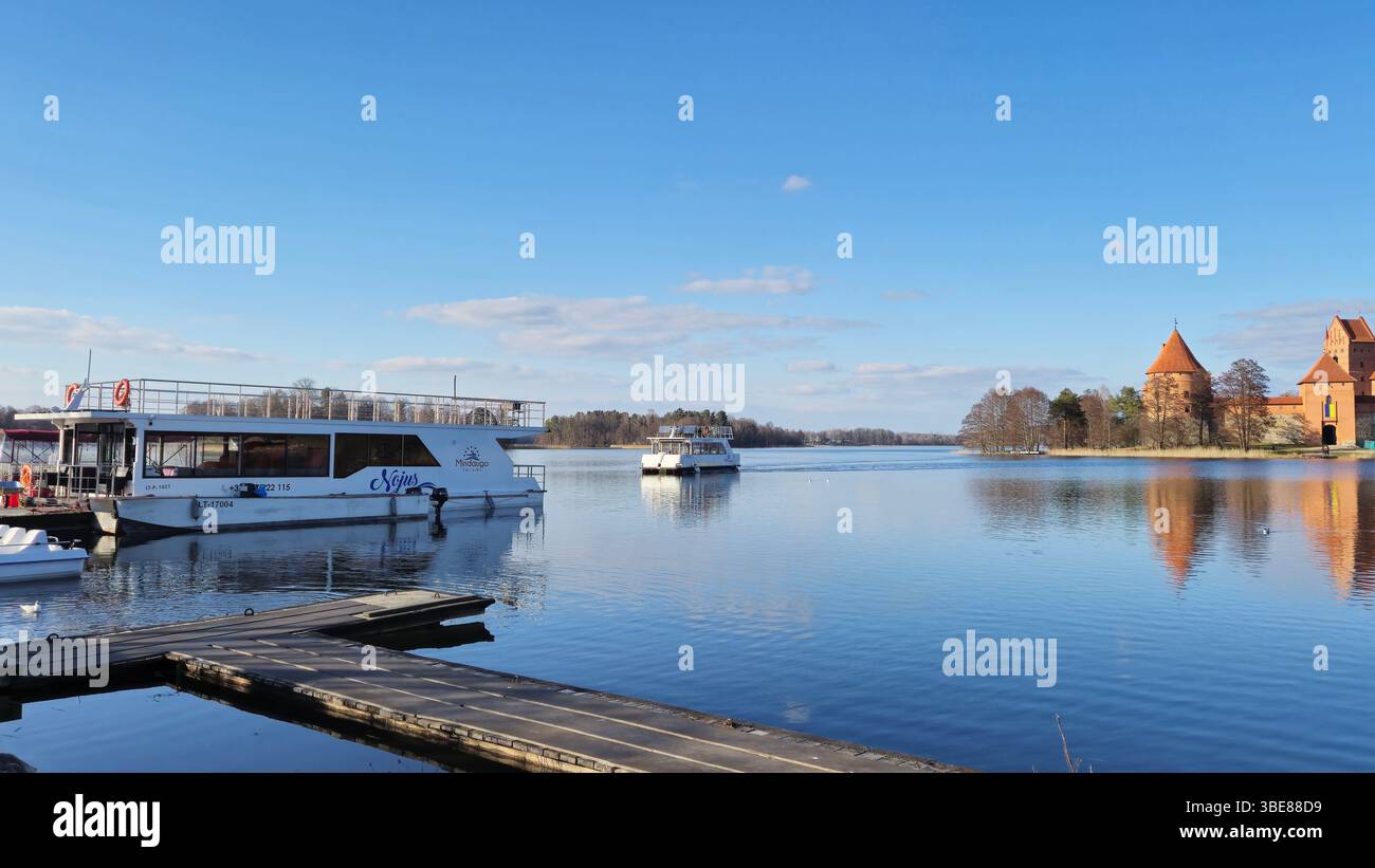 Trakai Lithuania - 03.28.2025 Tour boats on a calm lake with a view of a red brick medieval castle reflecting in the water under a clear blue sky - Smartphone Captured Stock Image