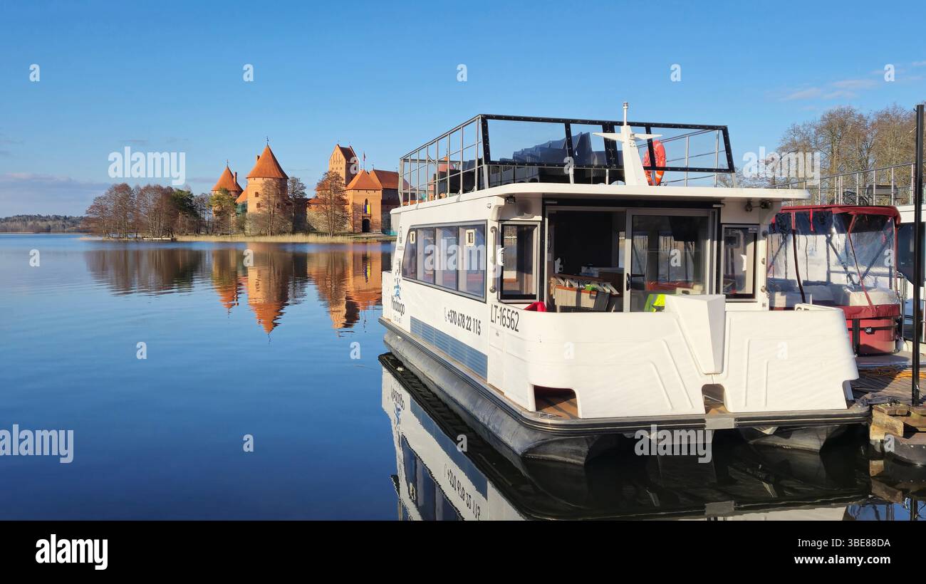 Trakai Lithuania - 03.28.2025 Tourist boat docked on tranquil lake with clear reflection of medieval castle and trees under blue sky on sunny day - Smartphone Captured Stock Image
