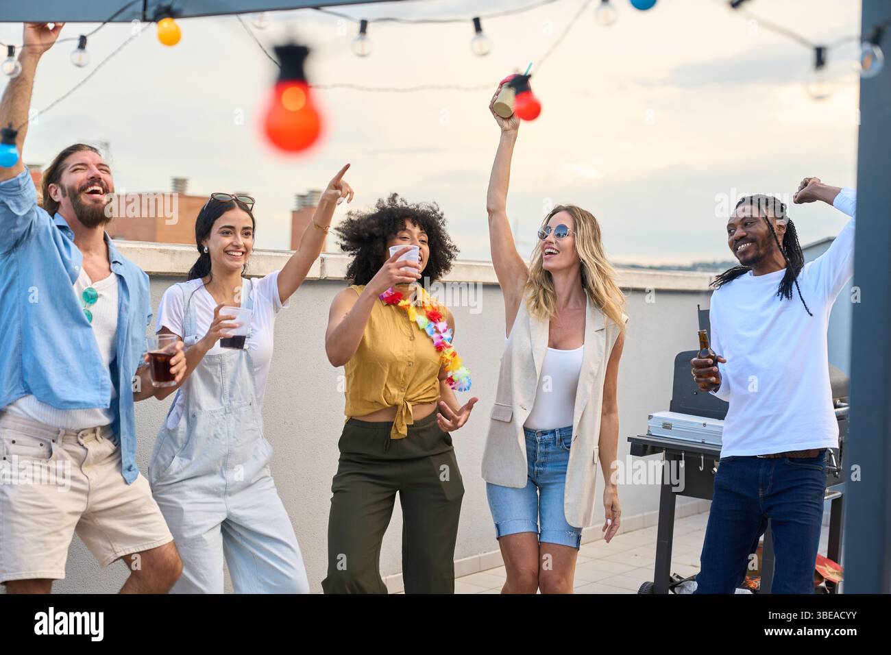 Friends dancing and drinking at rooftop party under string lights Stock Photo