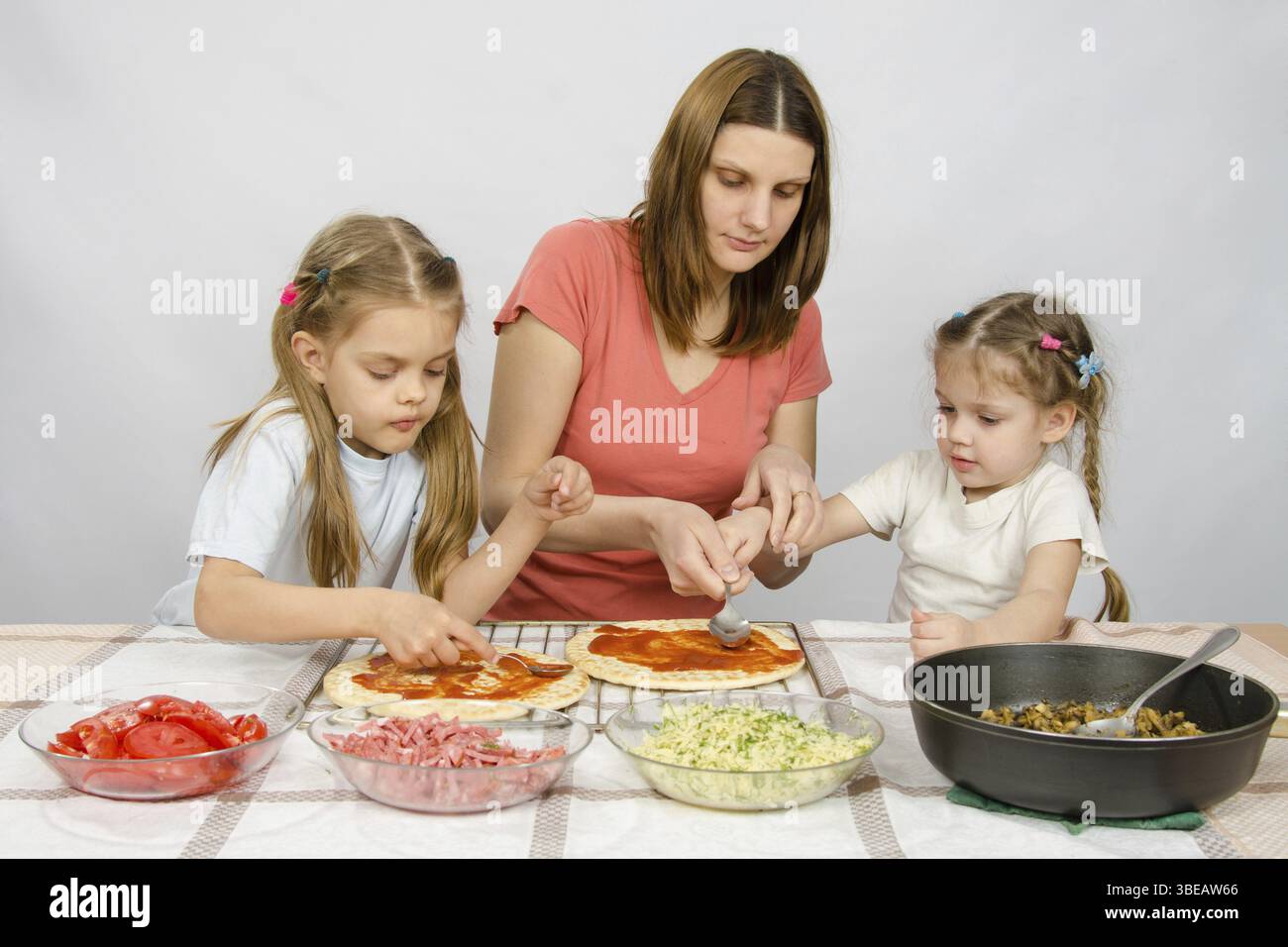 Mom helps younger daughter spread ketchup on a pizza, the eldest daughter, she is preparing a second pizza Stock Photo