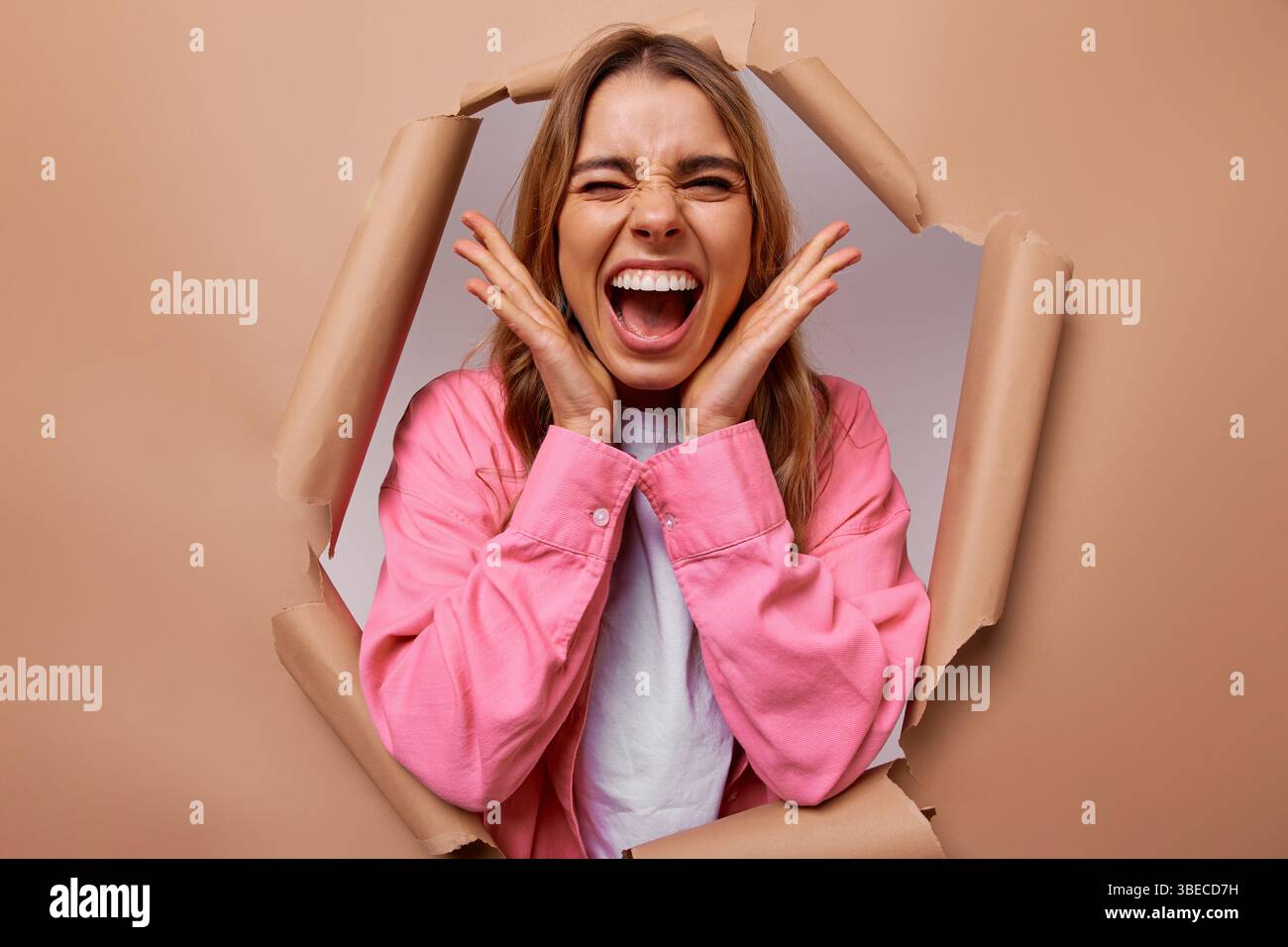 A vibrant young woman smiles widely and poses confidently, playfully peeking through a torn paper backdrop, embodying joy, creativity, and the delight Stock Photo