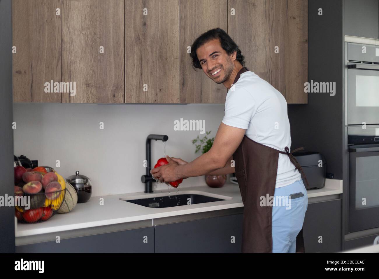 Man in apron washing a red bell pepper in the kitchen, smiling at the camera. Stock Photo