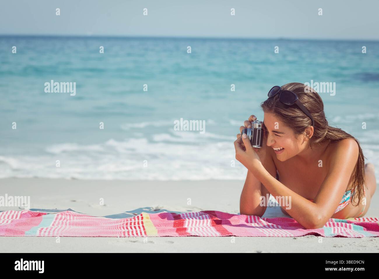 Woman in her twenties wearing floral bikini lying on beach towel, holding retro camera, copy space Stock Photo