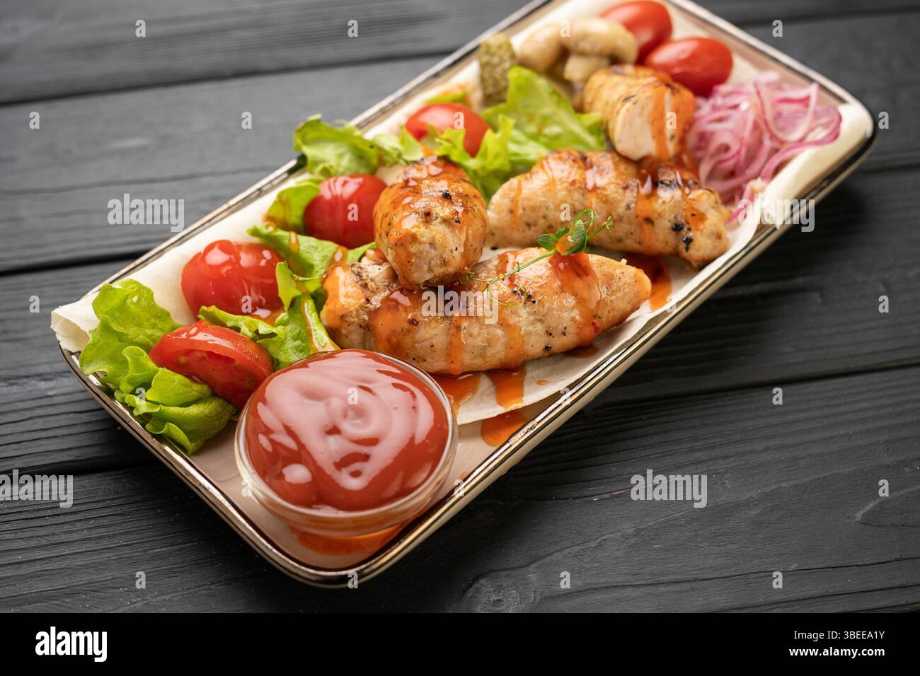 Juicy fried chicken kupaty served with ketchup, fresh tomatoes, and green lettuce on dark wooden table. German cuisine concept. Tasty homemade sausage Stock Photo