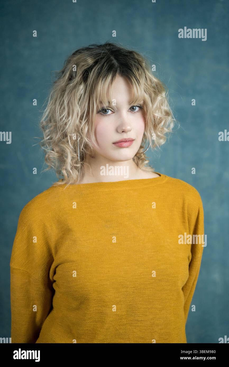 A young woman with curly blonde hair poses confidently in a yellow sweater against a textured blue backdrop, showcasing casual style and relaxed elega Stock Photo