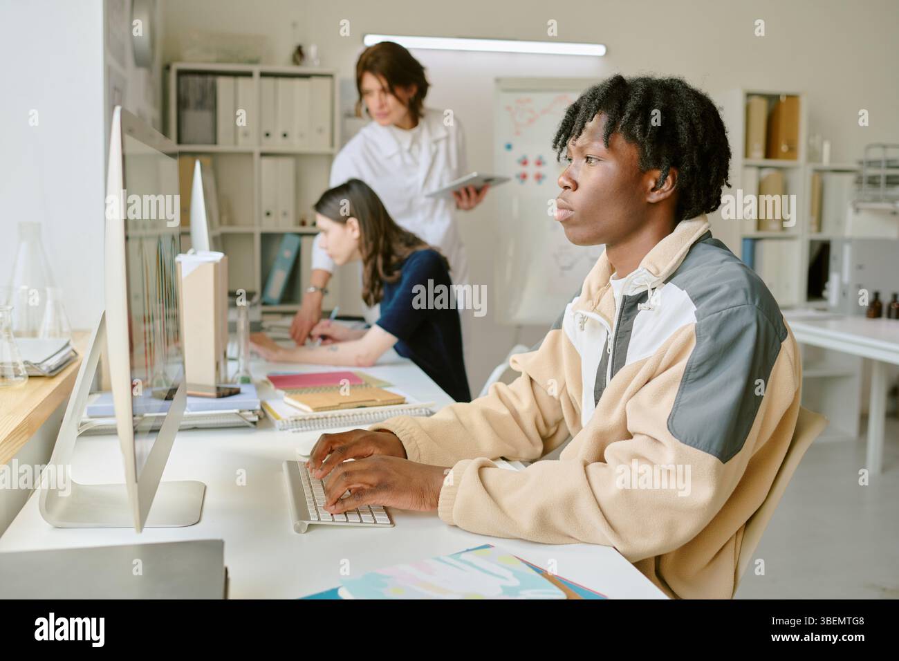 Student working on computer, focused and typing while two colleagues collaborate in background in bright, organized office space featuring various wor Stock Photo