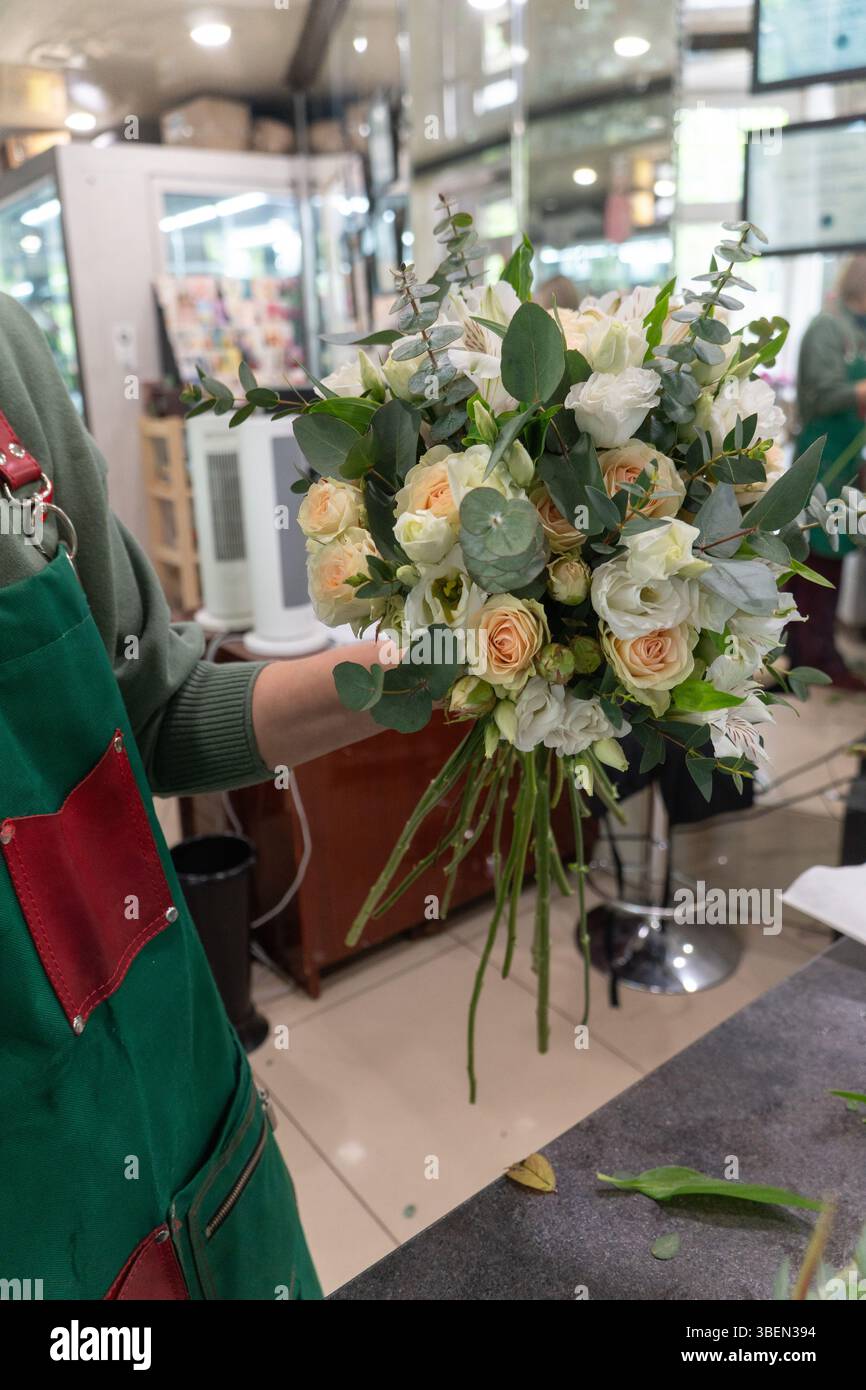 Florist holding a handcrafted bouquet with ivory and peach roses, lisianthus, and eucalyptus, in a well-lit flower shop. Artisanal floristry Stock Photo