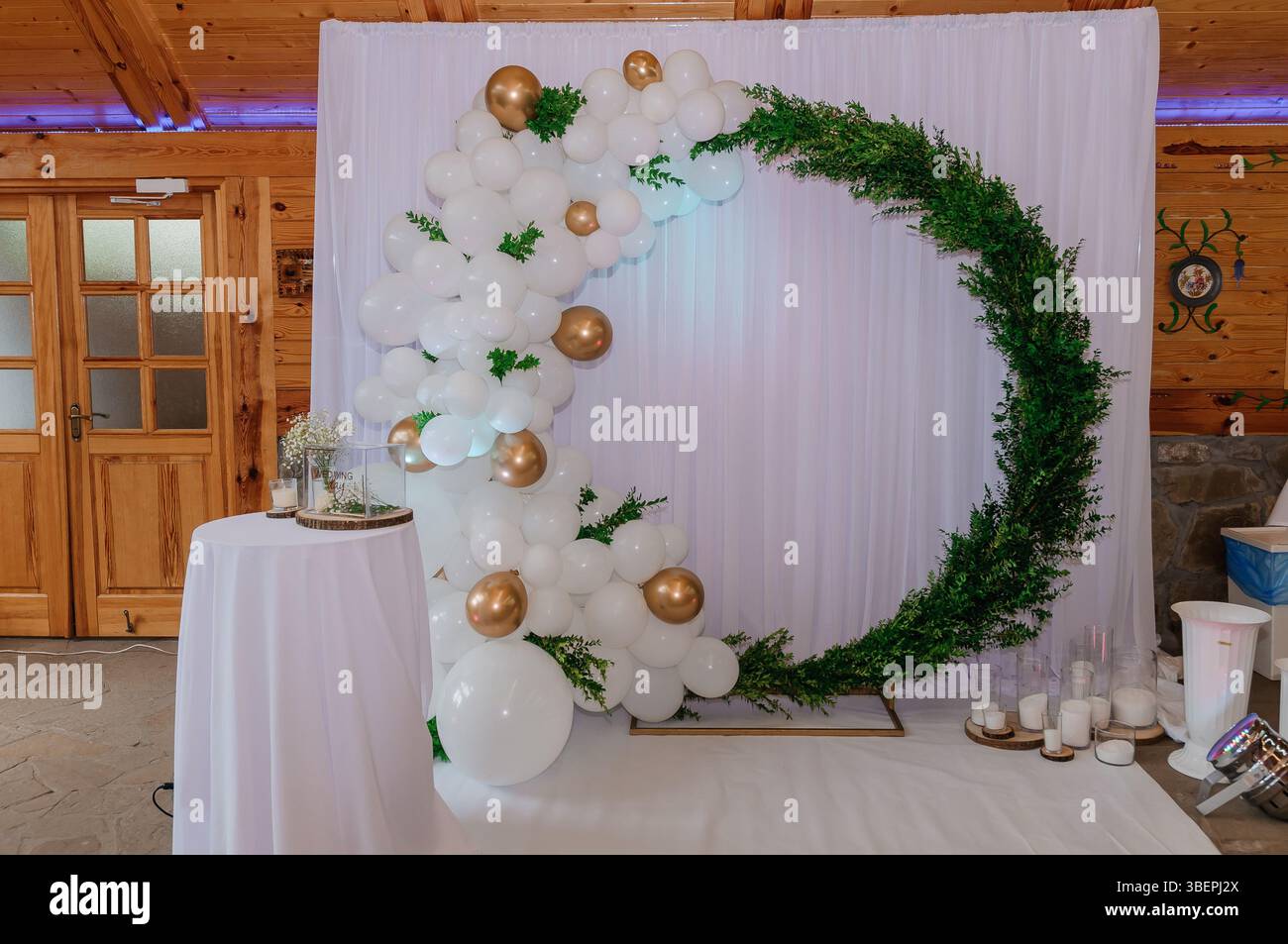 Elegant wedding backdrop featuring a white fabric drape adorned with a lush balloon garland in shades of white and gold, complemented by vibrant green Stock Photo