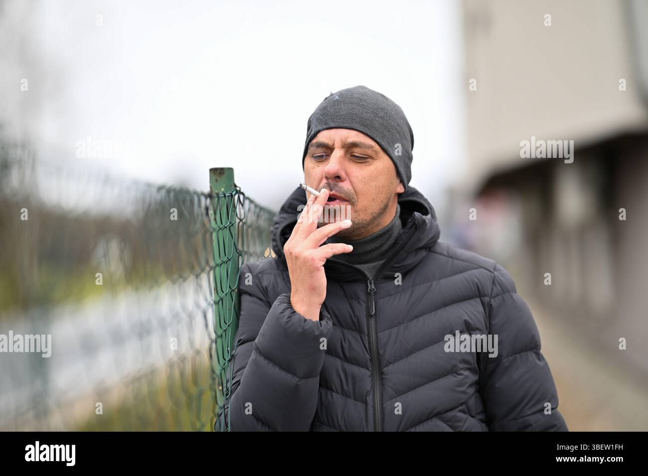 Man Smoking Outside.Man in winter jacket and beanie smoking a cigarette  near a chain link fence outdoors Stock Photo