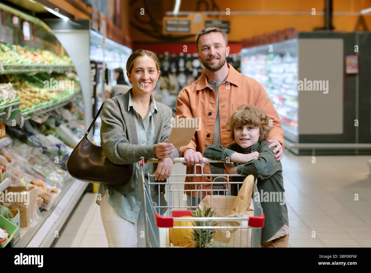 Family of three standing in grocery store, smiling while holding shopping list and cart with groceries. Capturing happy moment during routine grocery Stock Photo
