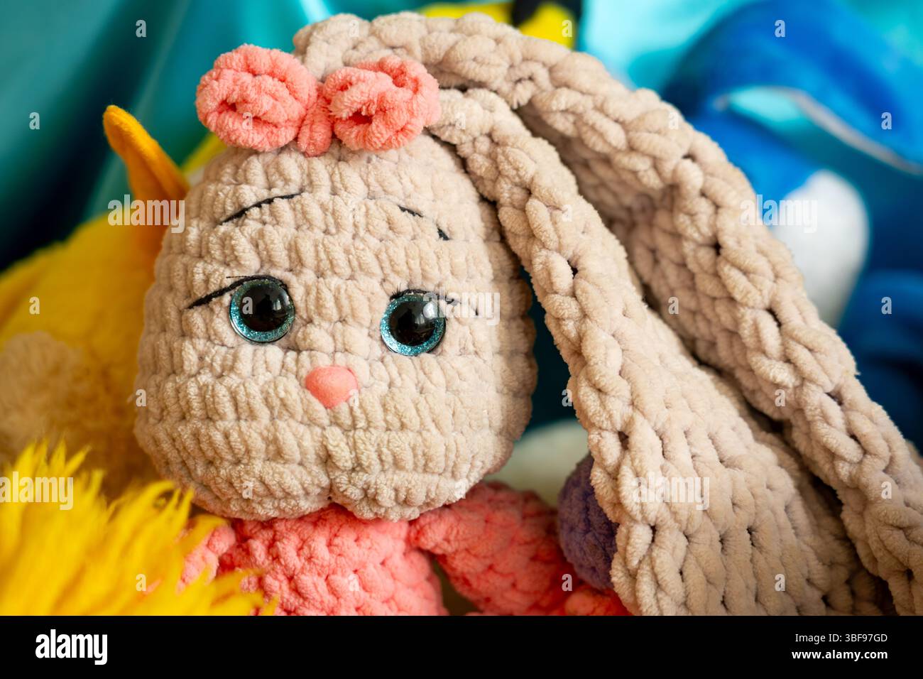 A cute plush bunny toy with fabric texture features big blue eyes and a pink bow. It is placed among other colorful stuffed animals in a cozy environm Stock Photo
