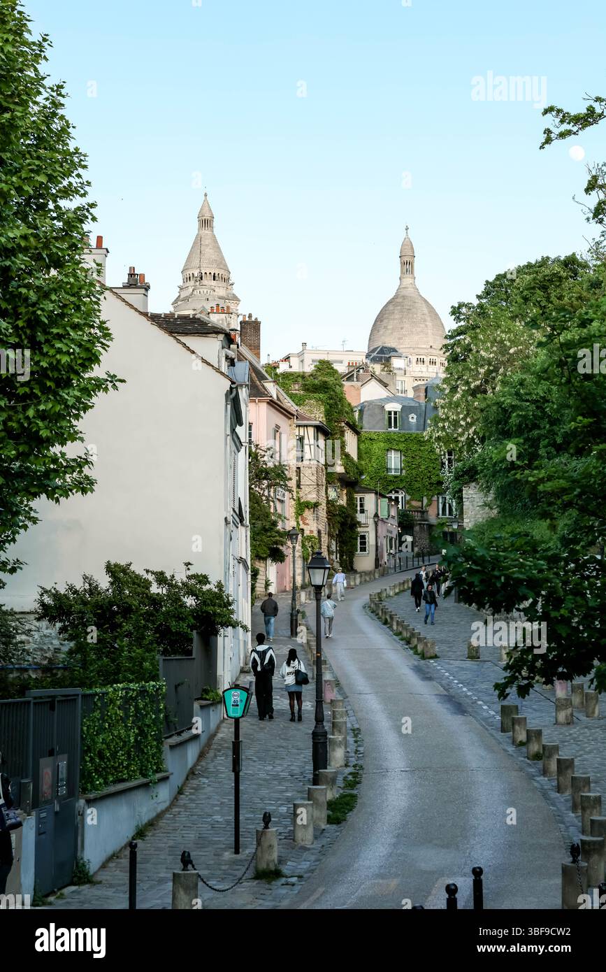 A classic Montmartre street scene looking uphill along a narrow, paved road towards the domes of the Sacré-Cœur Basilica, with pedestrians walking. Stock Photo