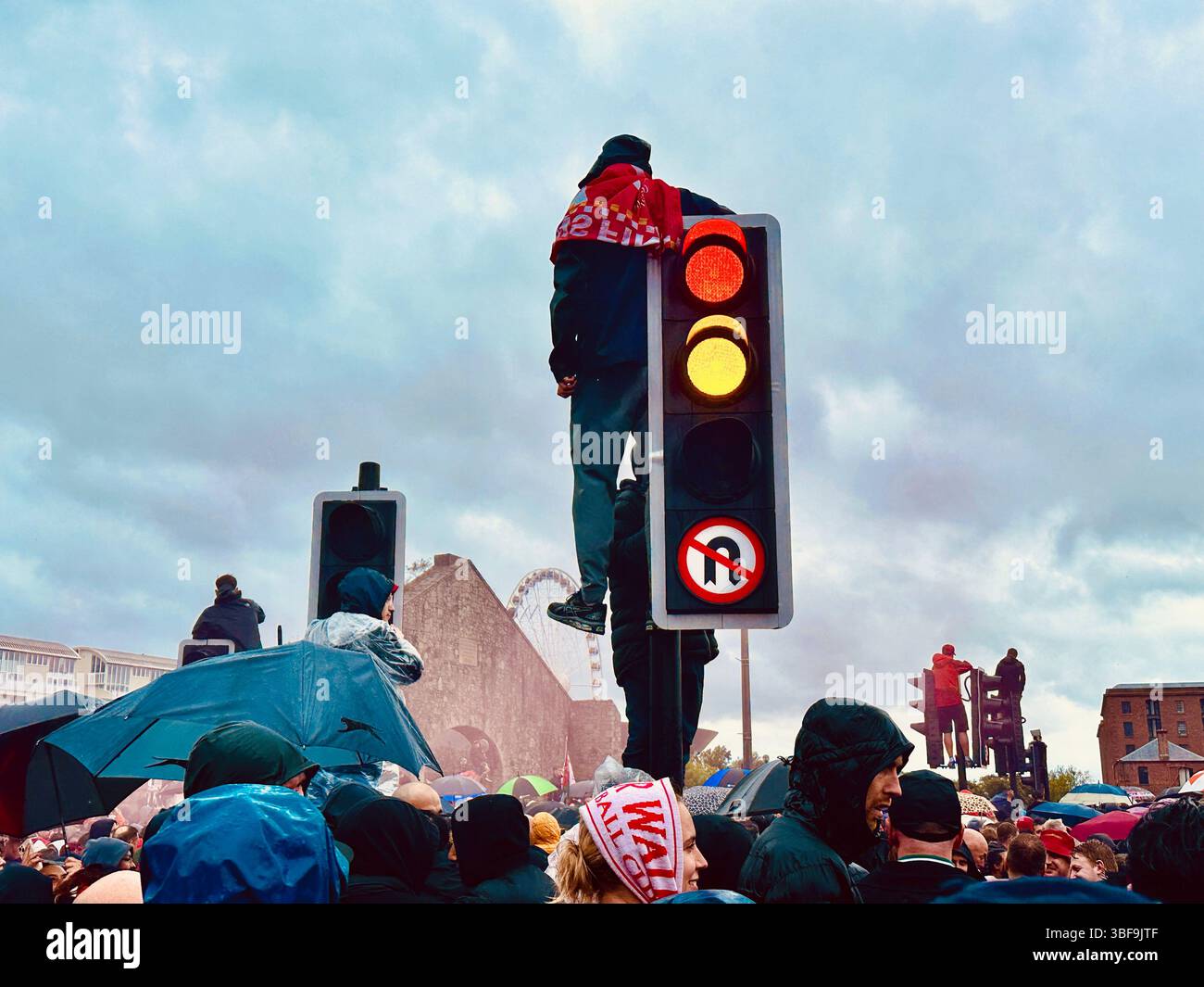 Liverpool fan with flag around shoulders on traffic light above crowd on The Strand during victory parade, with red smoke and supporters, 25 May 2025 - Smartphone Captured Stock Image