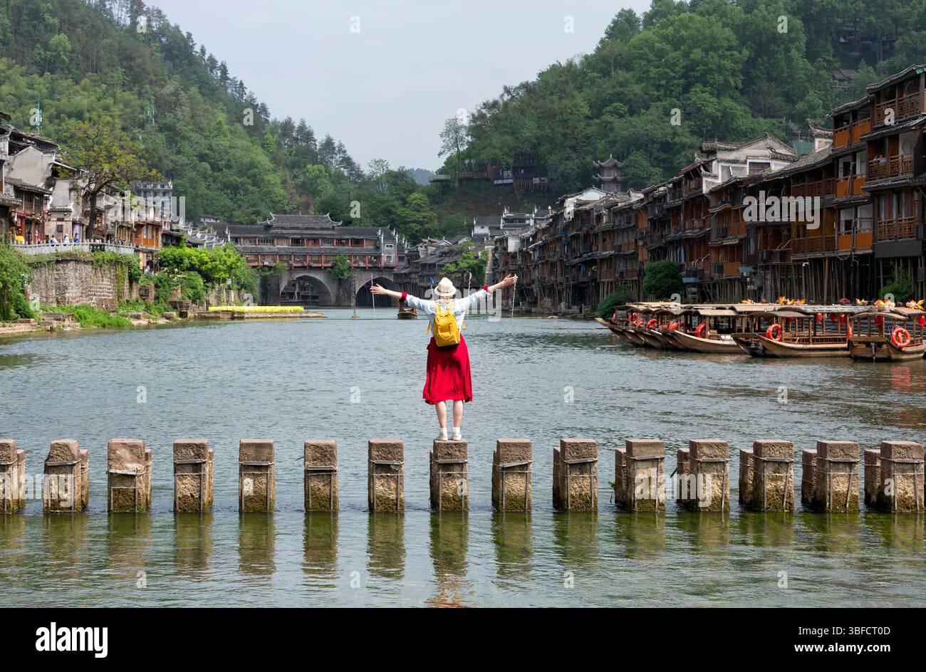 Young tourist dressed in red crossing the Tuojiang River in the historic Xiangxi Phoenix Ancient Town or Fenghuang Ancient Old Town, Hunan, China. Stock Photo