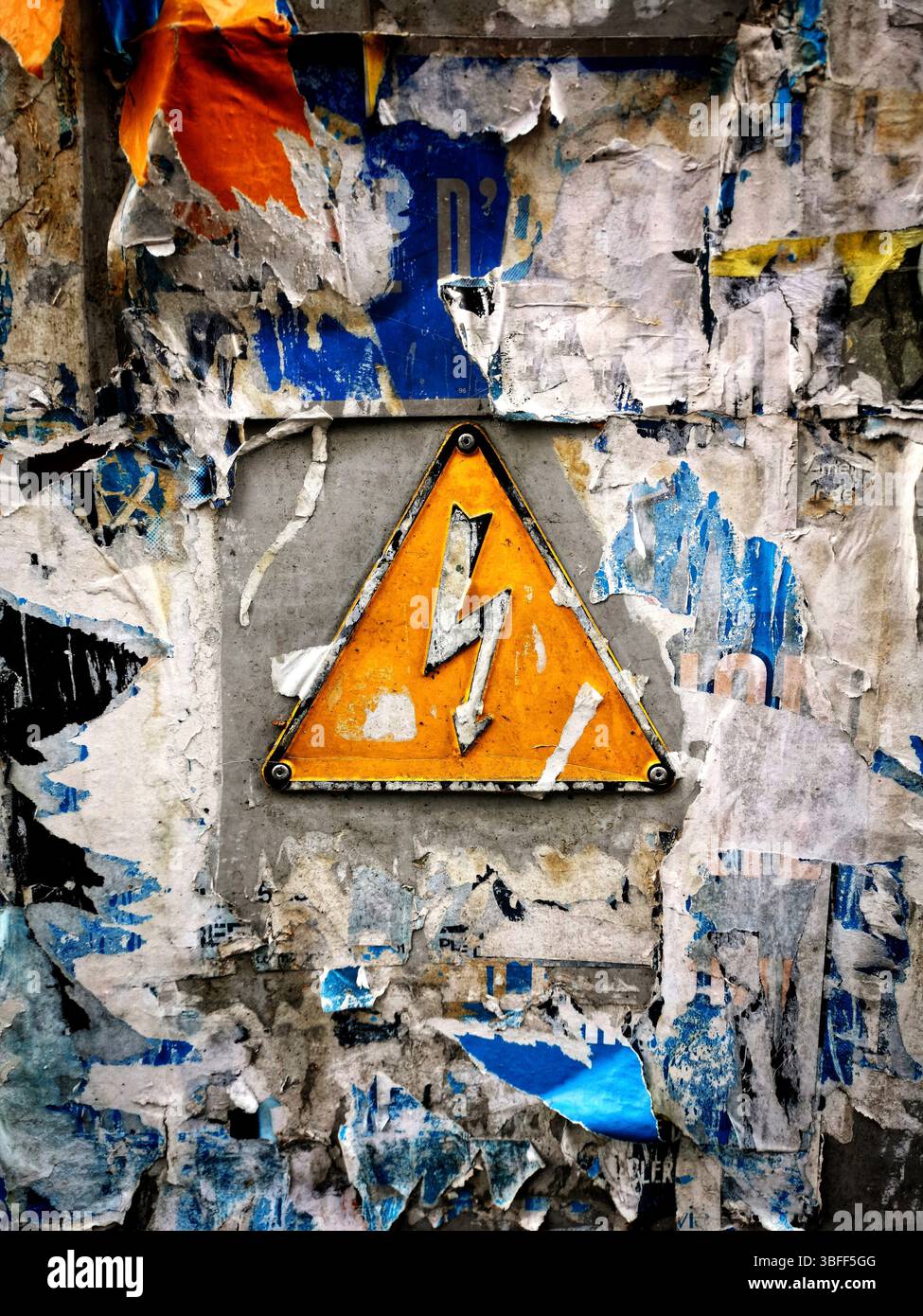 A bright yellow warning sign featuring a lightning bolt is mounted on a wall heavily covered with faded, torn posters. - Smartphone Captured Stock Image