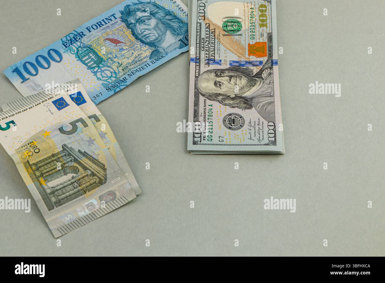 Various currency notes are displayed on a gray surface. A hundred-dollar bill sits next to a blue 1000 forint note and a five-euro note, showcasing in Stock Photo