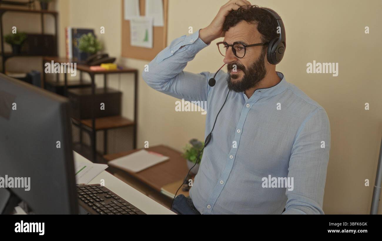 Hispanic man with beard wearing headset in office setting, engaging through computer for virtual communication, emphasizing modern workspace atmospher Stock Photo