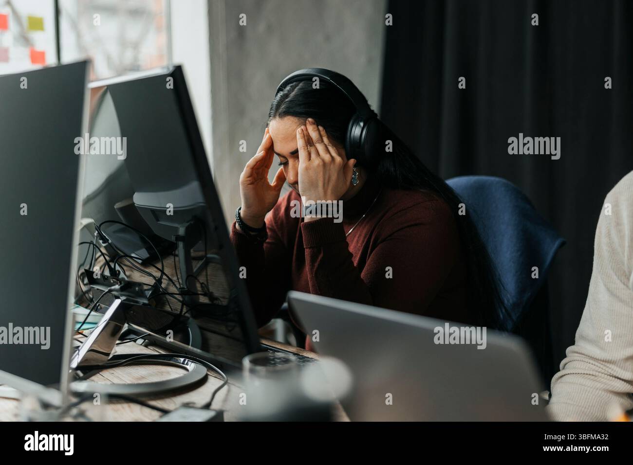 Exhausted female programmer touching temples while sitting at desk in tech start up office Stock Photo