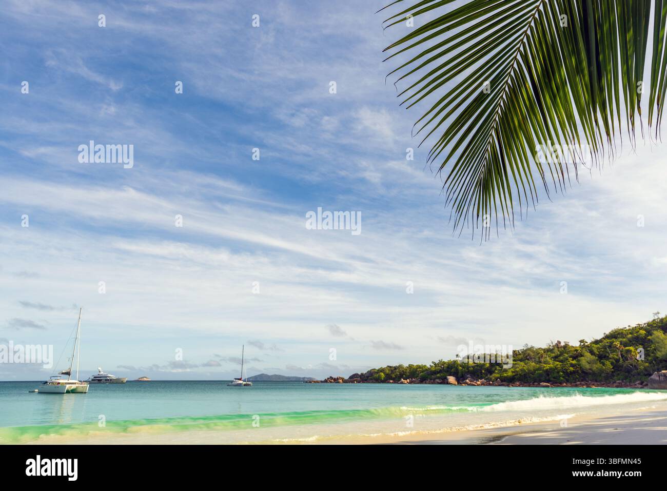 A serene tropical beach scene featuring turquoise waters, anchored sailboats, and lush greenery. Highlights include a clear sky, a palm leaf in the fo Stock Photo
