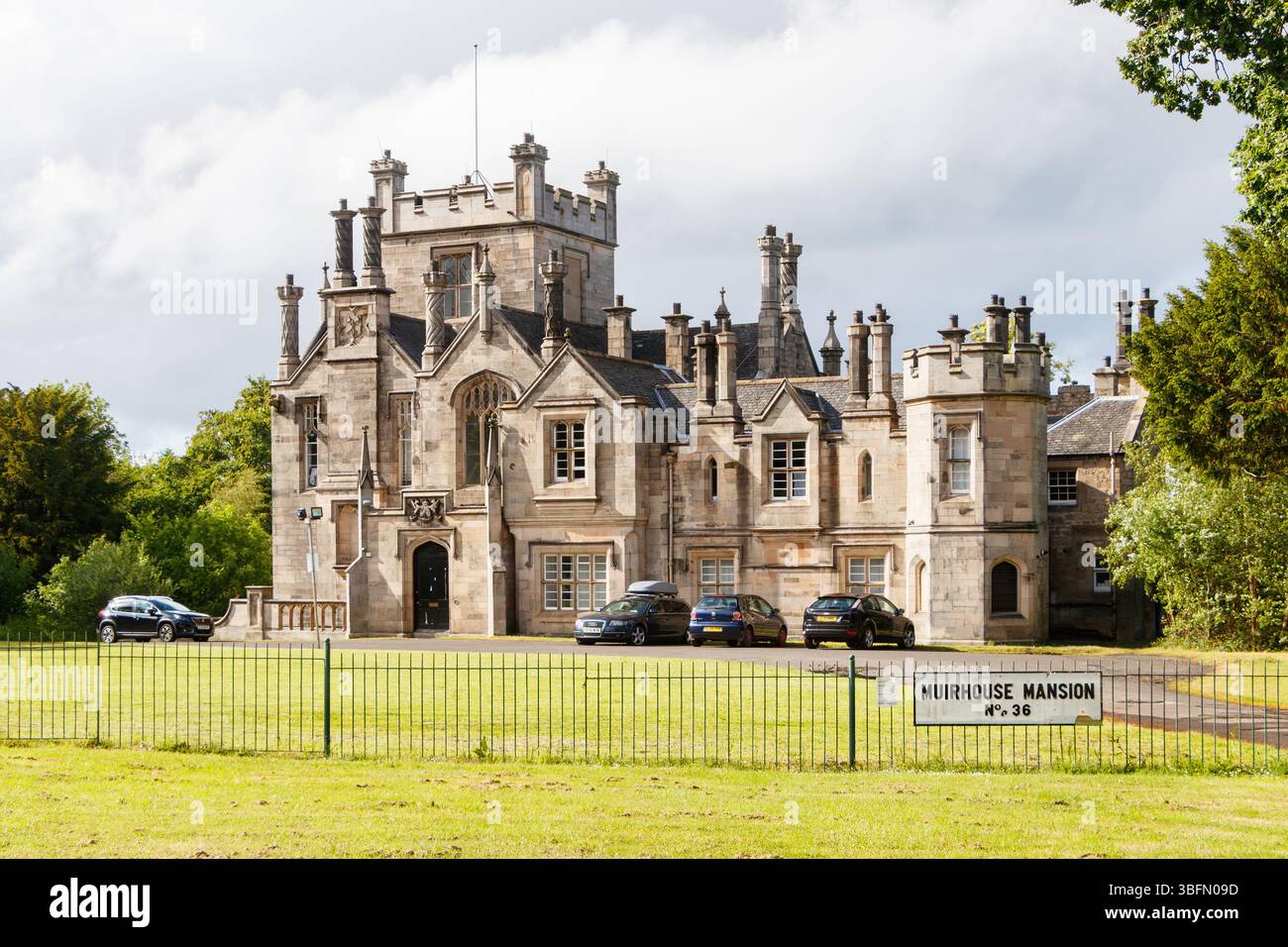 Historic stone mansion with turrets and chimneys surrounded by green lawn and parked cars, labeled Muirhouse Mansion No. 36 Stock Photo