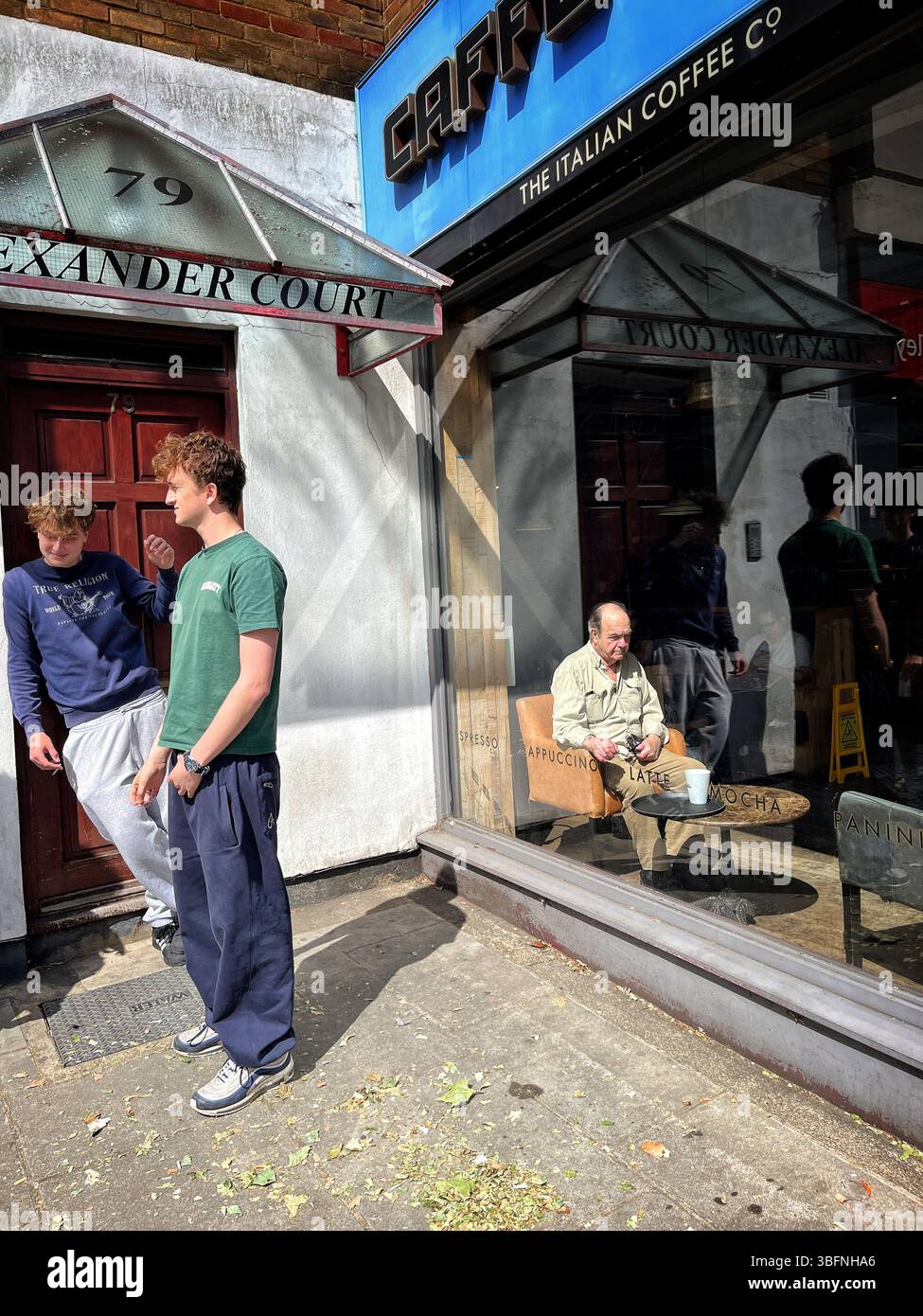 Two youths, who look like twins, stand outside a cafe, whilst inside the coffee shop, an elderly bald man sits at a table alone. - Smartphone Captured Stock Image