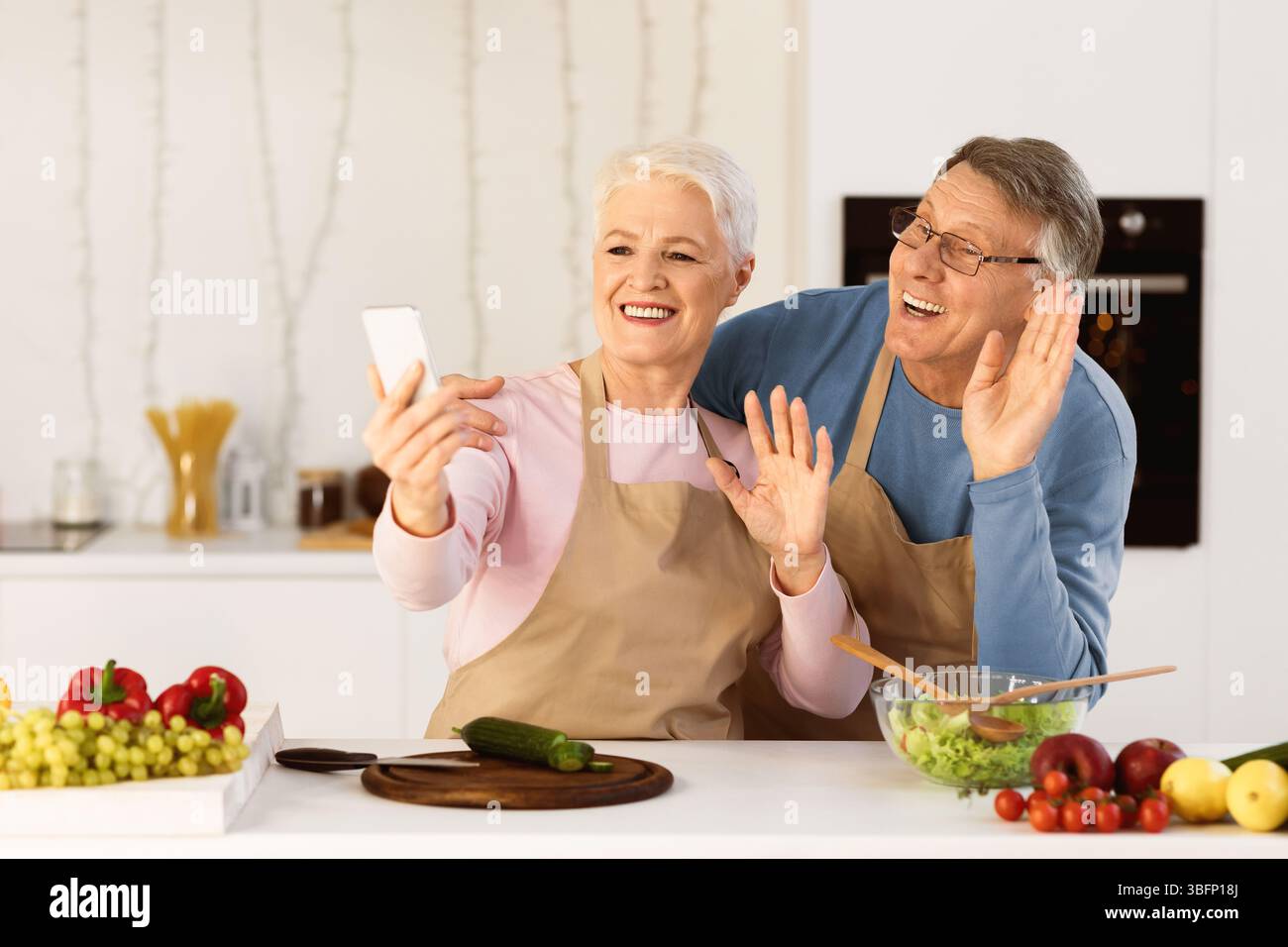 Senior Couple Making Video Call Using Mobile Phone In Kitchen Stock Photo