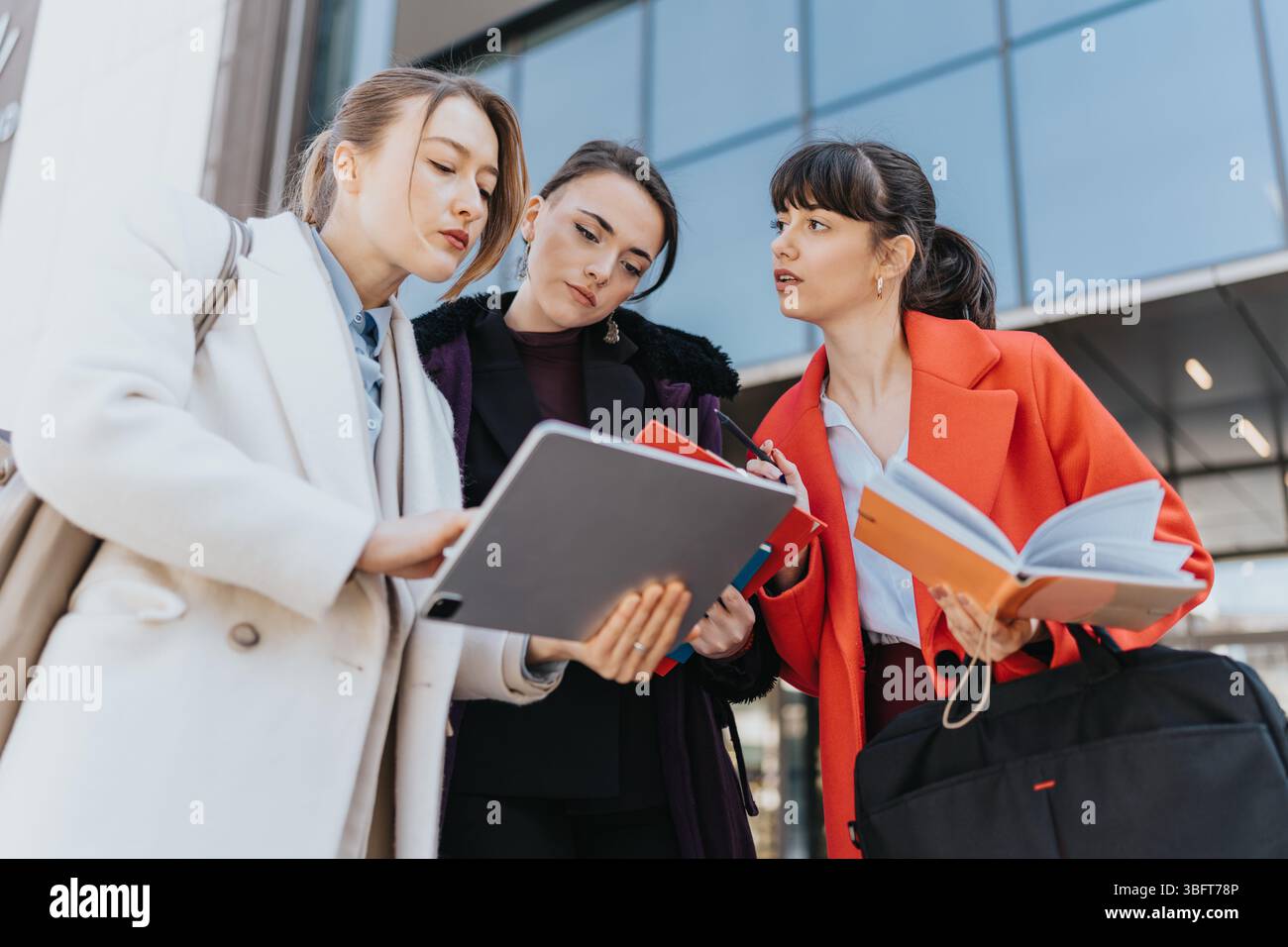 Three businesswomen collaborating outdoors while reviewing notes and using digital devices Stock Photo