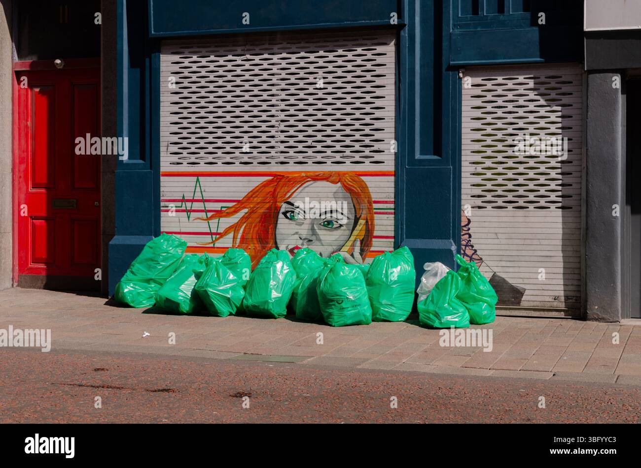 Belfast County Antrim Northern Ireland April 09 2025 - Shop shutter with a woman's head painted on it obscured by green rubbish bags Stock Photo
