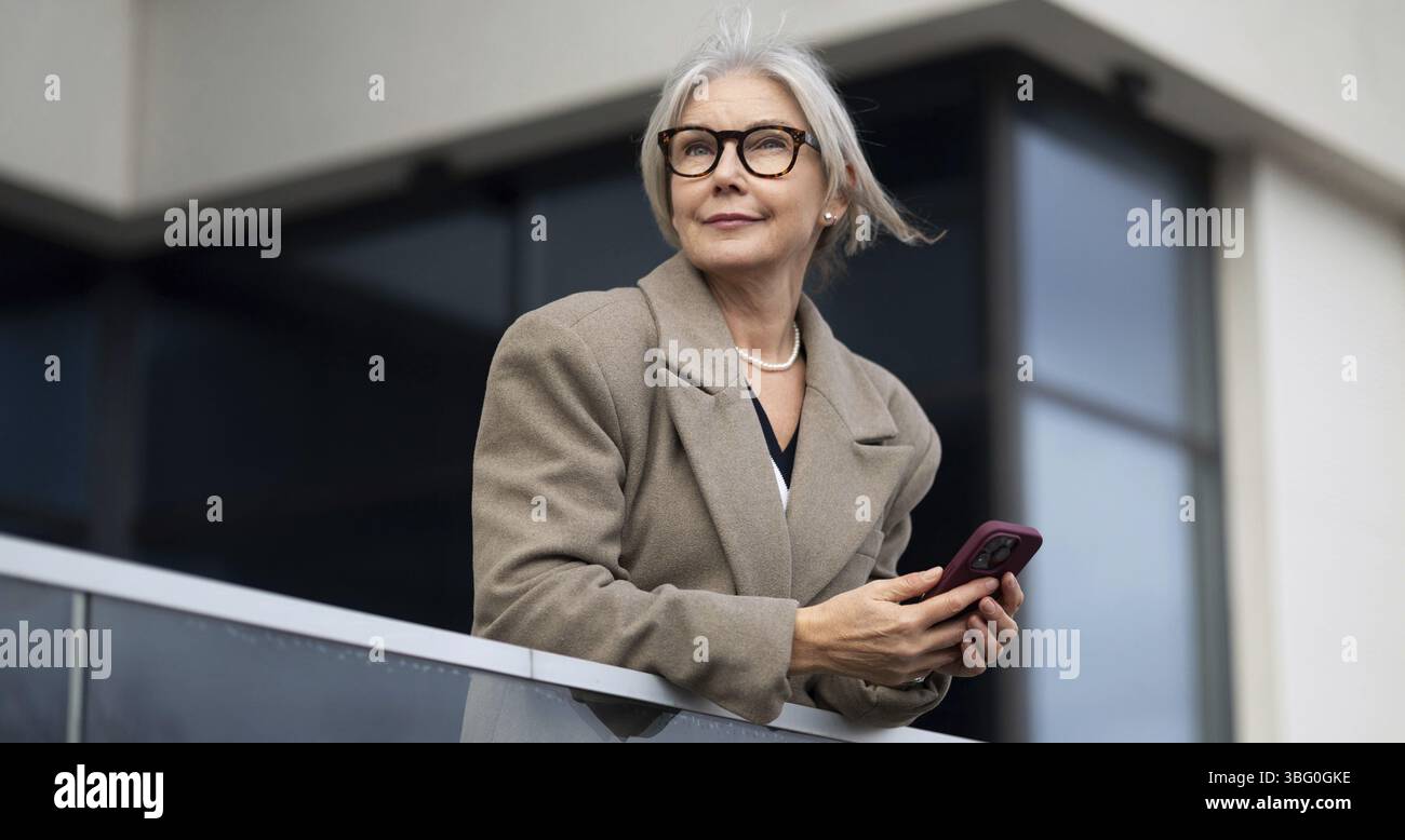 The stylish elderly woman stands elegantly on a balcony, holding her phone and gazing thoughtfully into the distance, bathed in warm afternoon light Stock Photo