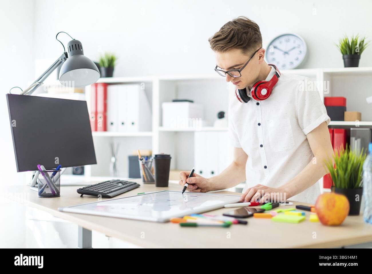 A young man in glasses stands near a computer desk. A young man draws a ...