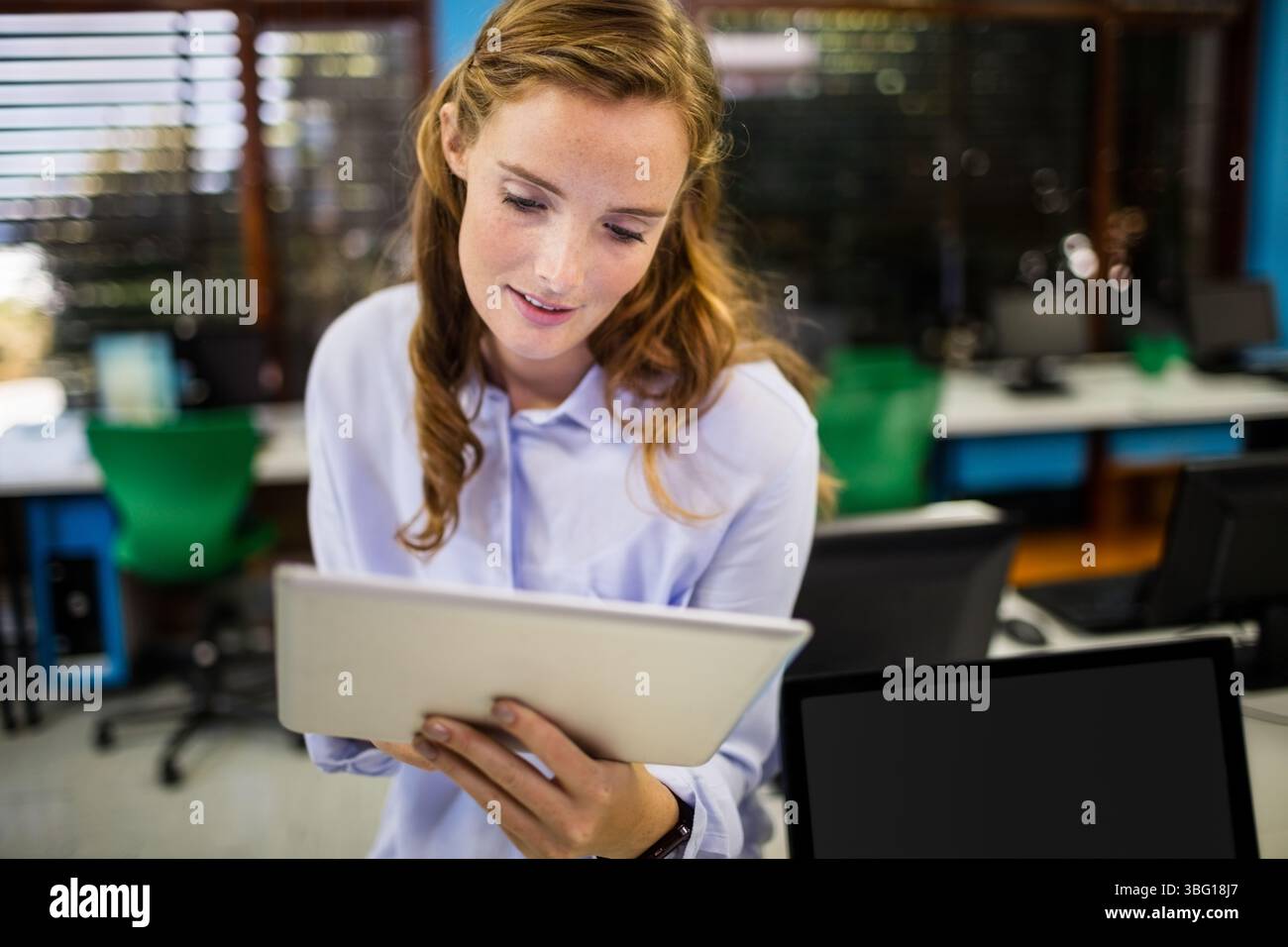 Tablet displaying content on desk in computer lab with monitors green chairs and wooden blinds Stock Photo
