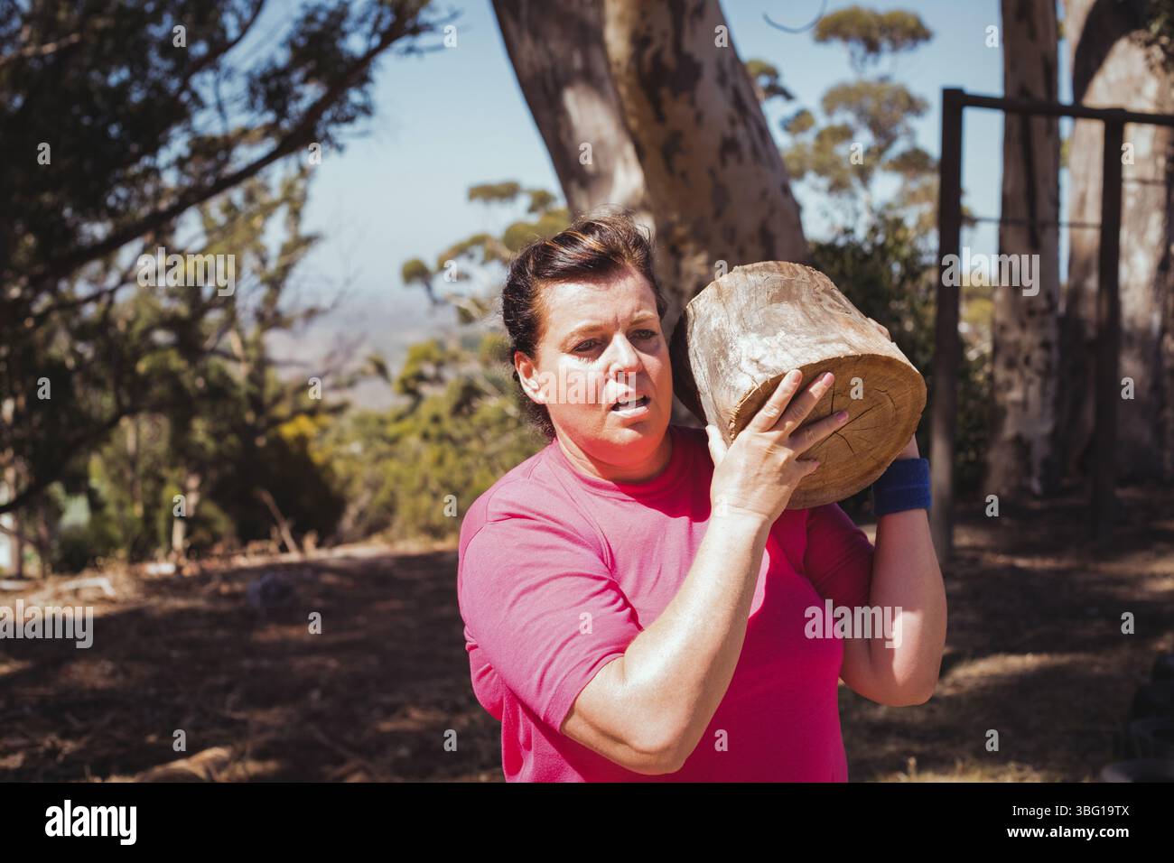Woman lifting wooden log in sunlit forest clearing wearing pink T-shirt and blue wristband Stock Photo
