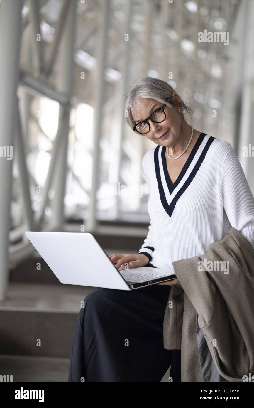 An older woman, dressed in a stylish sweater, is focused on her laptop in a bright, contemporary environment. She holds a coat and looks engaged in he Stock Photo