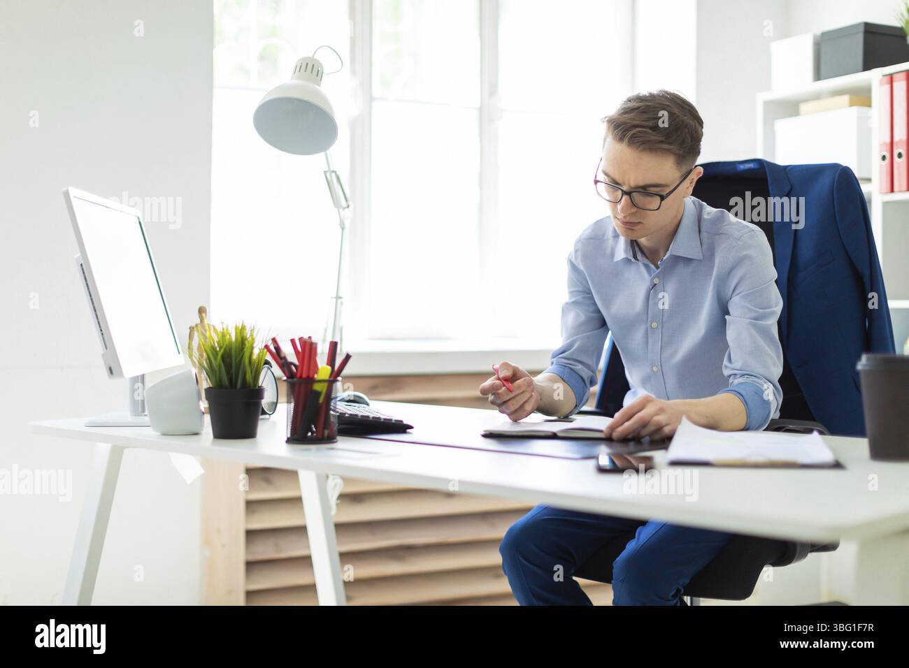 A handsome young man with glasses, blue trousers and a light shirt is working in the office. On the back of the chair hangs his jacket. photo with dep Stock Photo