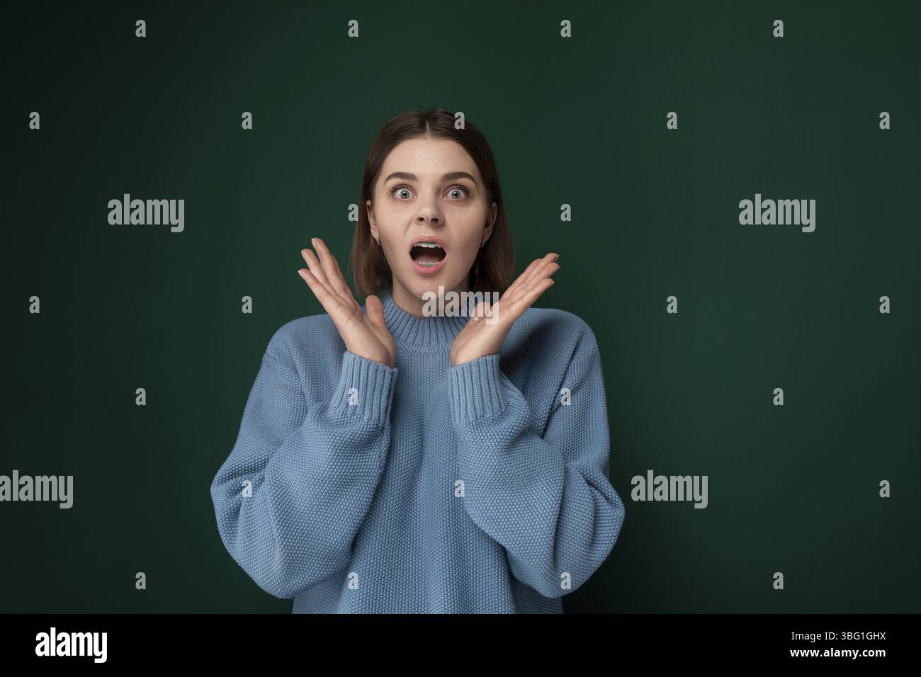 A woman wearing a blue sweater is shown making a facial expression indicative of surprise. Her eyes are widened and mouth slightly agape Stock Photo