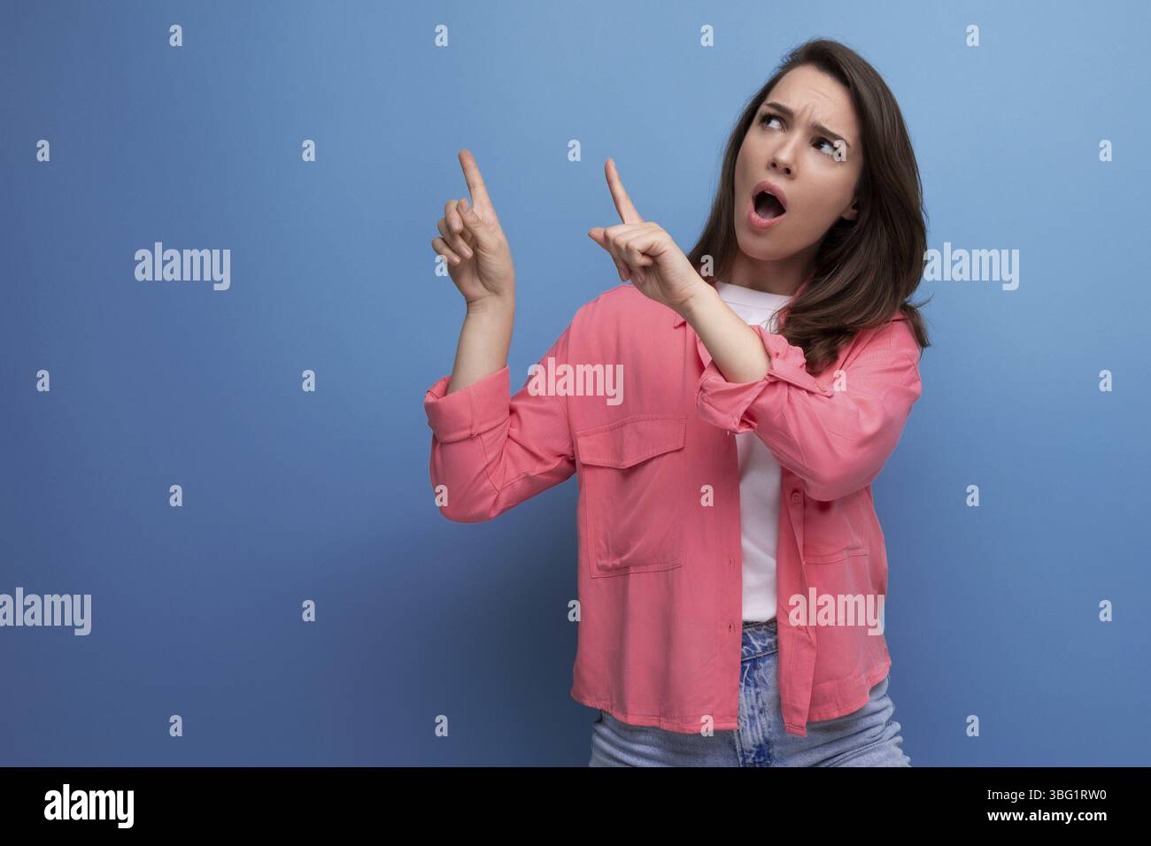 Shocked black haired lady in casual style pointing finger at wall in studio background Stock Photo