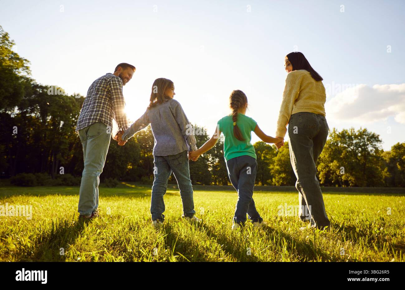 Back view of father, mother, daughter son holding hands together standing in fiend on green grass Stock Photo