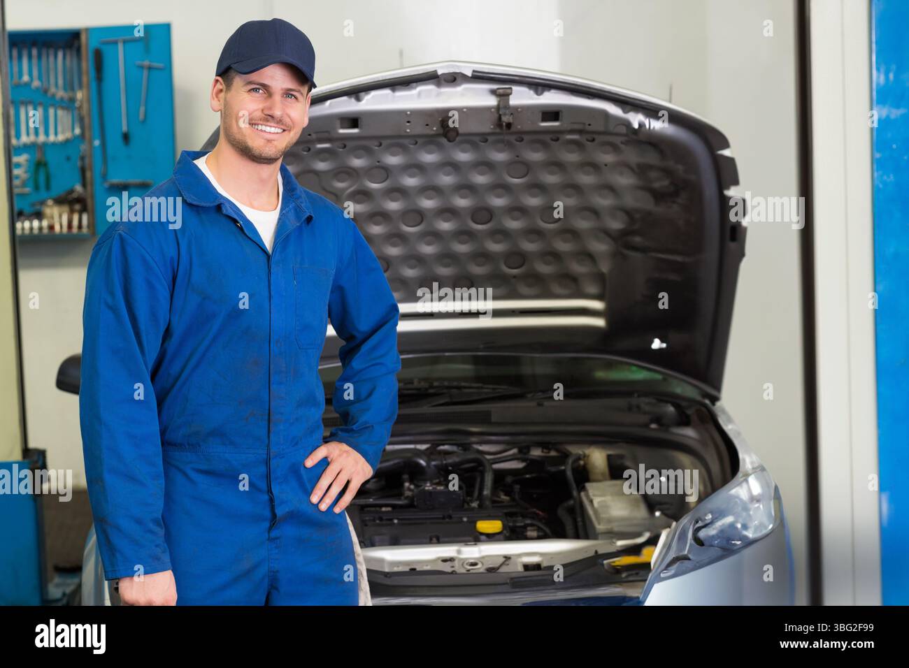 Male mechanic standing beside silver car with open hood at garage showing engine, tools, copy space Stock Photo