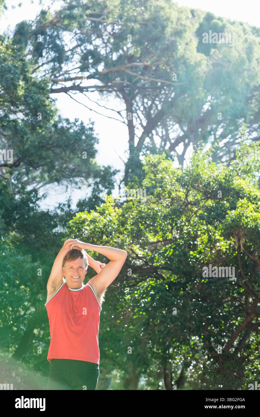Mature man in red top and black shorts stretching triceps raising arm overhead in sunlit woods Stock Photo