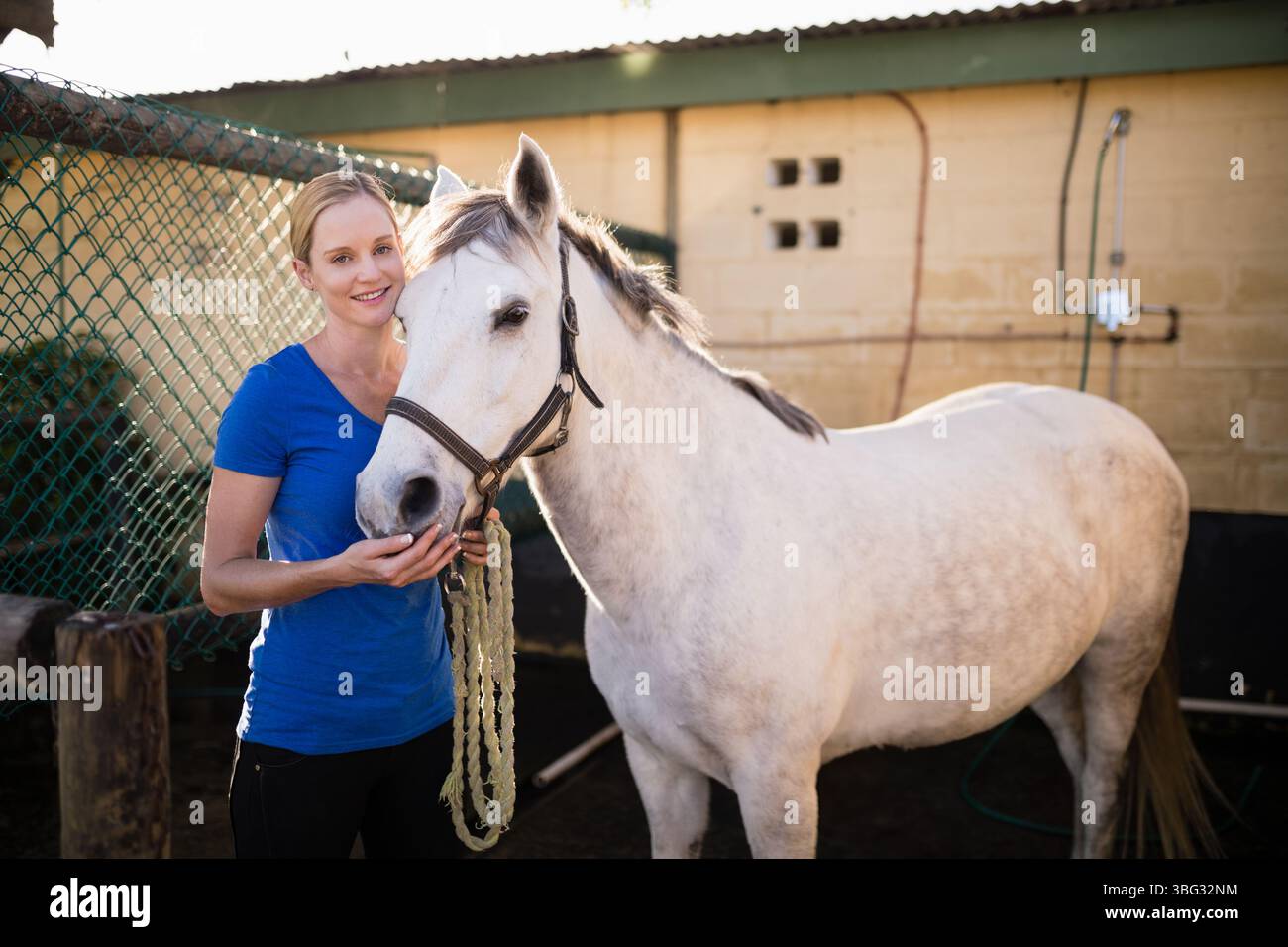 Woman holding braided lead rope touching white horse muzzle in stable ...