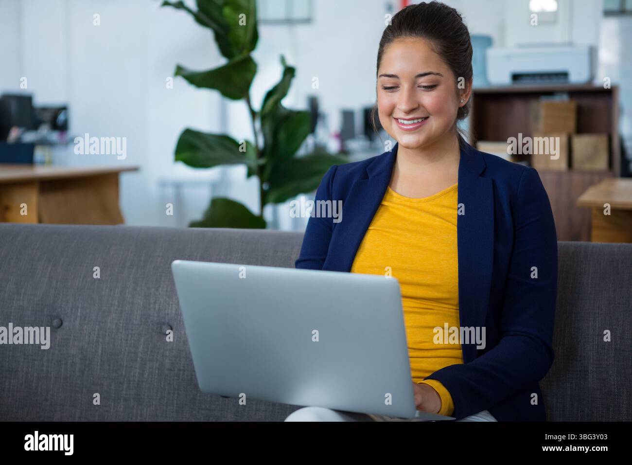 Woman working on laptop while sitting on gray sofa in modern office ...
