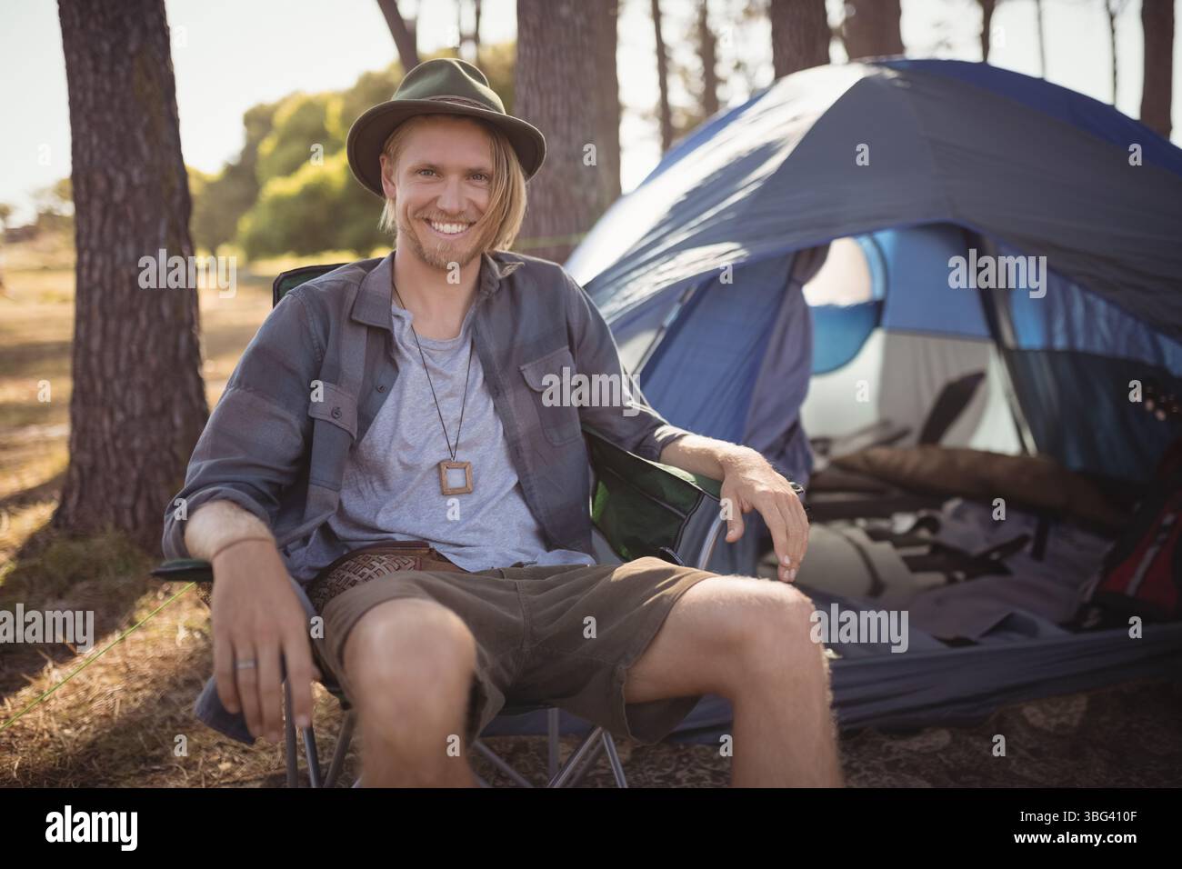 Male camper relaxing in collapsible camping chair at forest clearing with dome tent and backpack Stock Photo