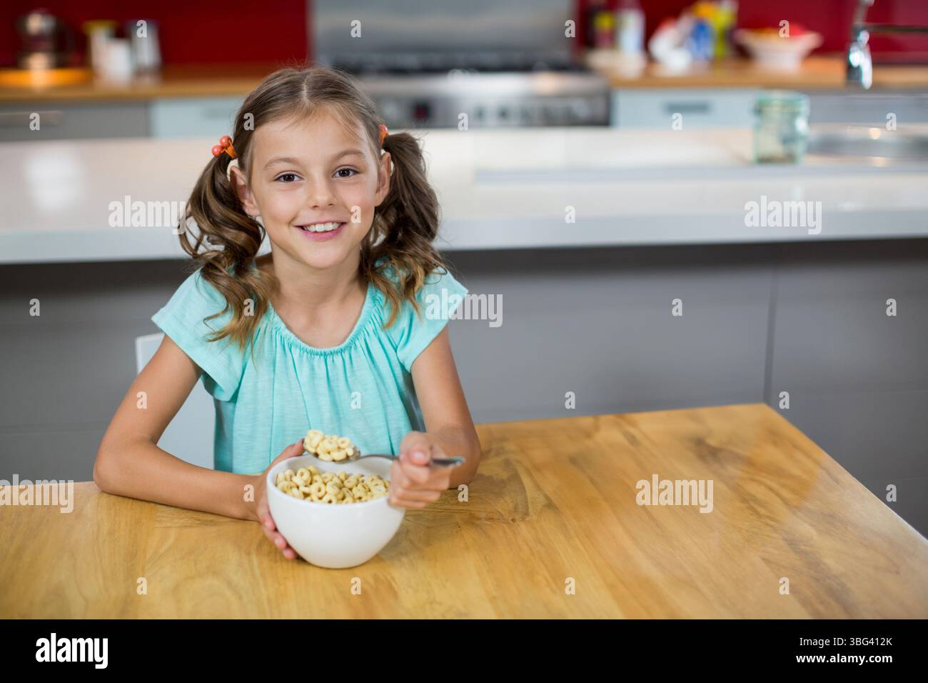 School-age girl holding metal spoon over white bowl and eating cereal at modern kitchen table Stock Photo